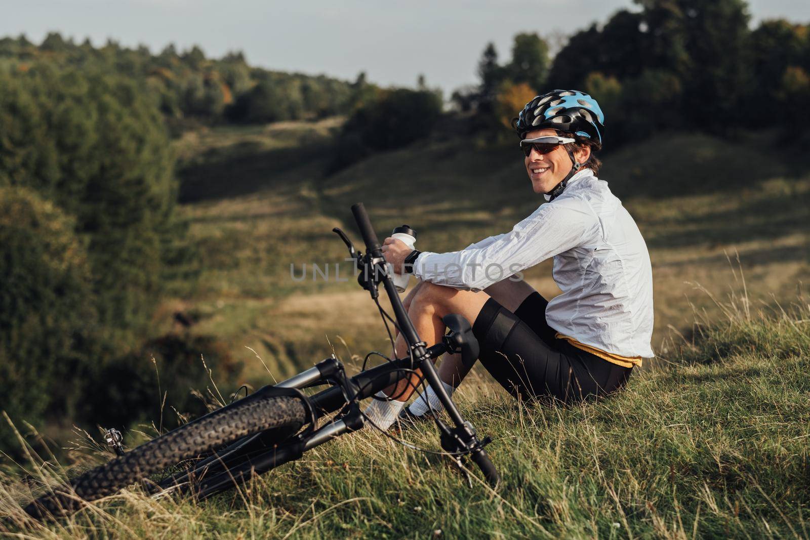 Professional Male Cyclist Drinking Water from Bottle, Man Sitting Near Bicycle During His Journey Outdoors in Countryside