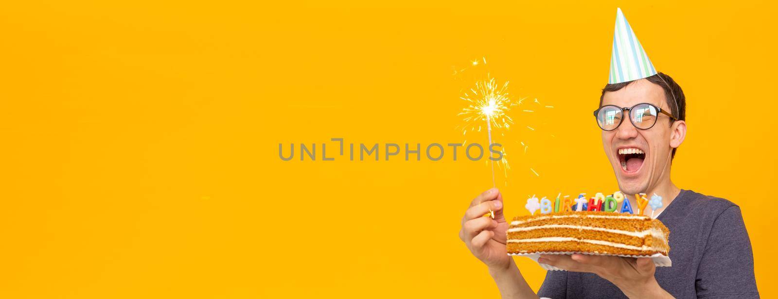 Banner funny positive guy in glasses holds in his hands a homemade cake with the inscription happy birthday posing on a yellow background. Concept of holidays and anniversaries. by Satura86