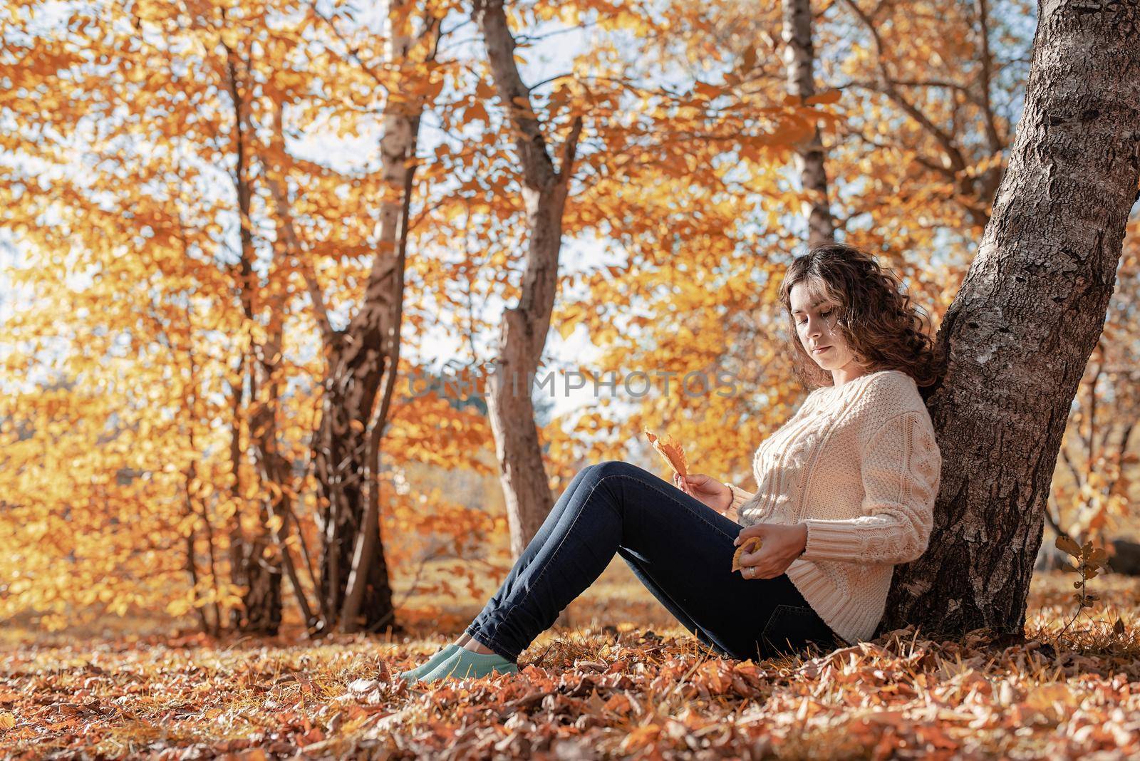 Autumn nature. Young thoughtful woman sitting by the tree in autumn forest