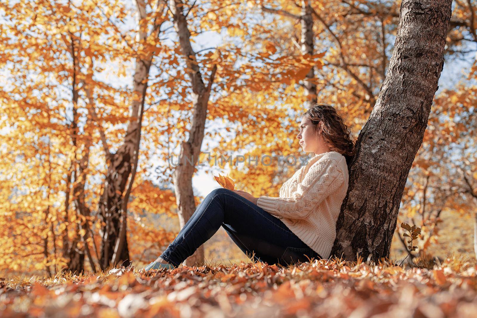 Autumn nature. Young thoughtful woman sitting by the tree in autumn forest