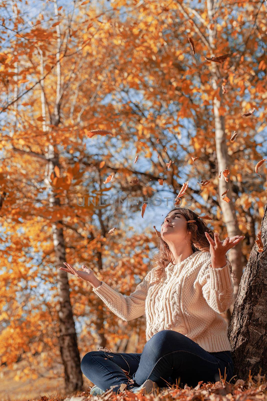 Autumn nature. Happy young woman throwing yellow leaves up in the air sitting in the autumn forest