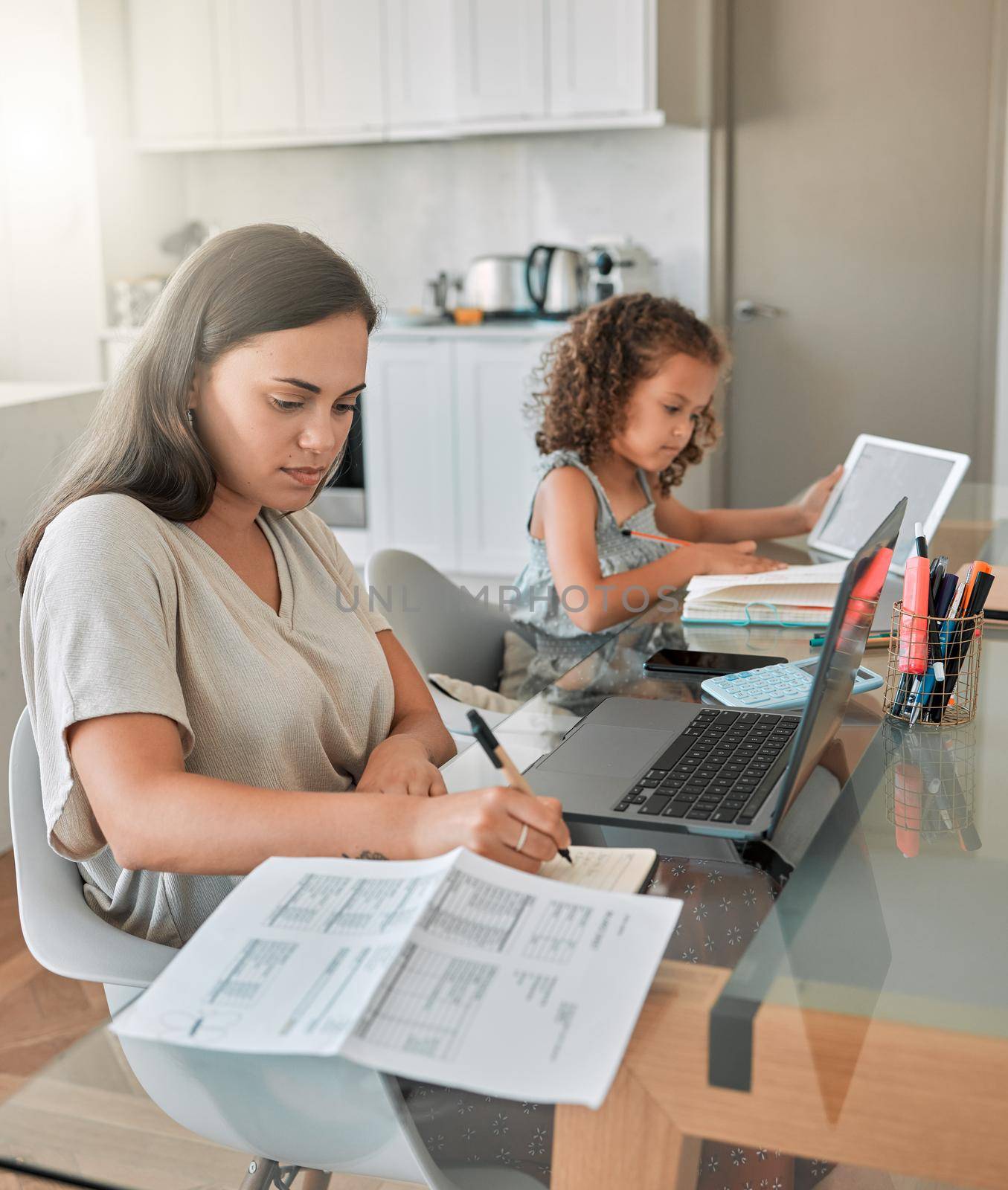 Mother and daughter being productive with remote work and homework, multitasking at a kitchen table at home. Parent and child serious while paying bills and watching an online education programme by YuriArcurs