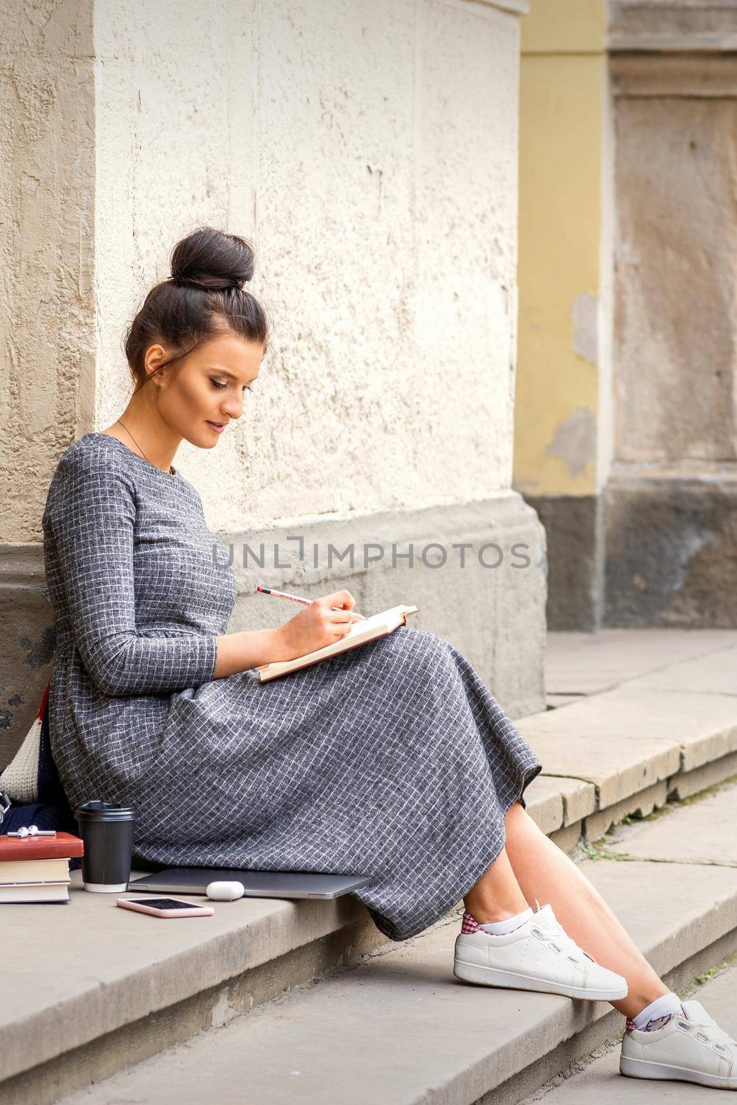 Female student in long dress reading a book on stairs of university building outdoors