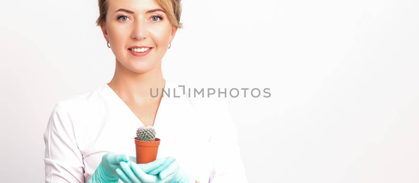 Young beautiful smiling female beautician holding a small cactus in a pot on white background