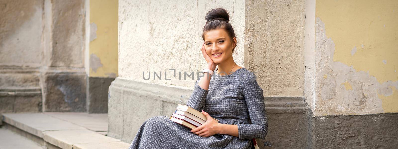 Female student in a long dress holding books looking at camera sitting on stairs of university building outdoors