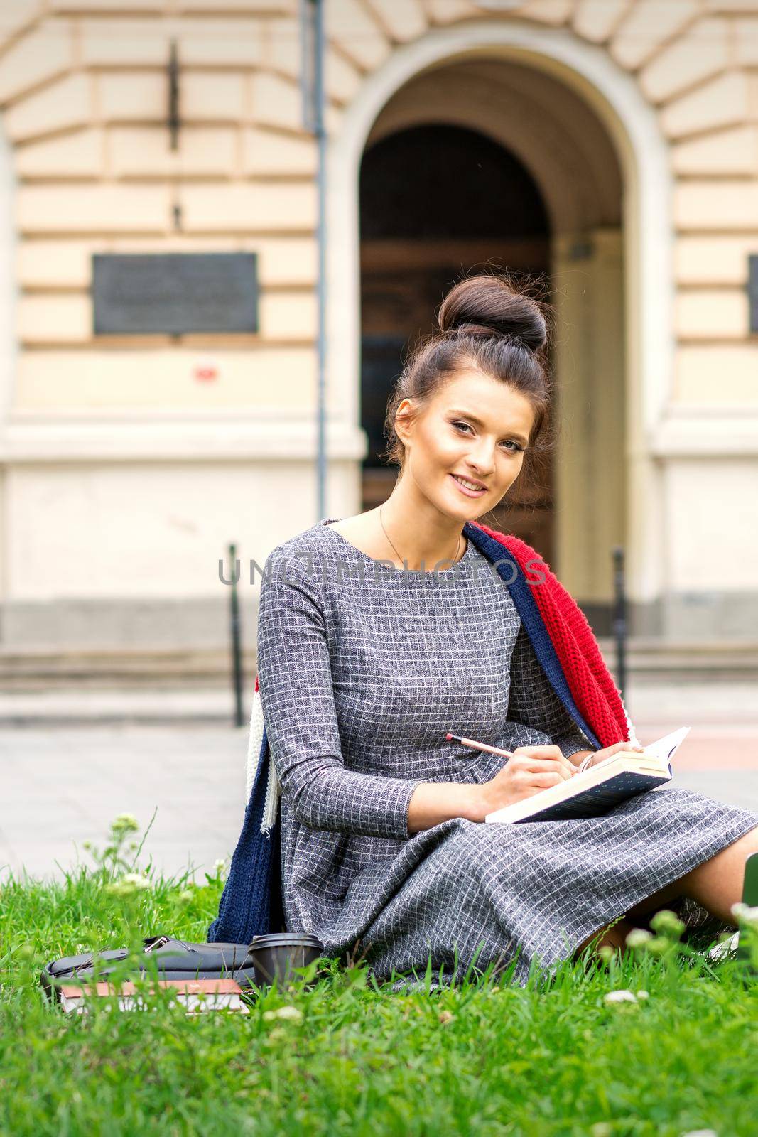 Portrait of a beautiful young smiling student studying with a book on the grass near the university building