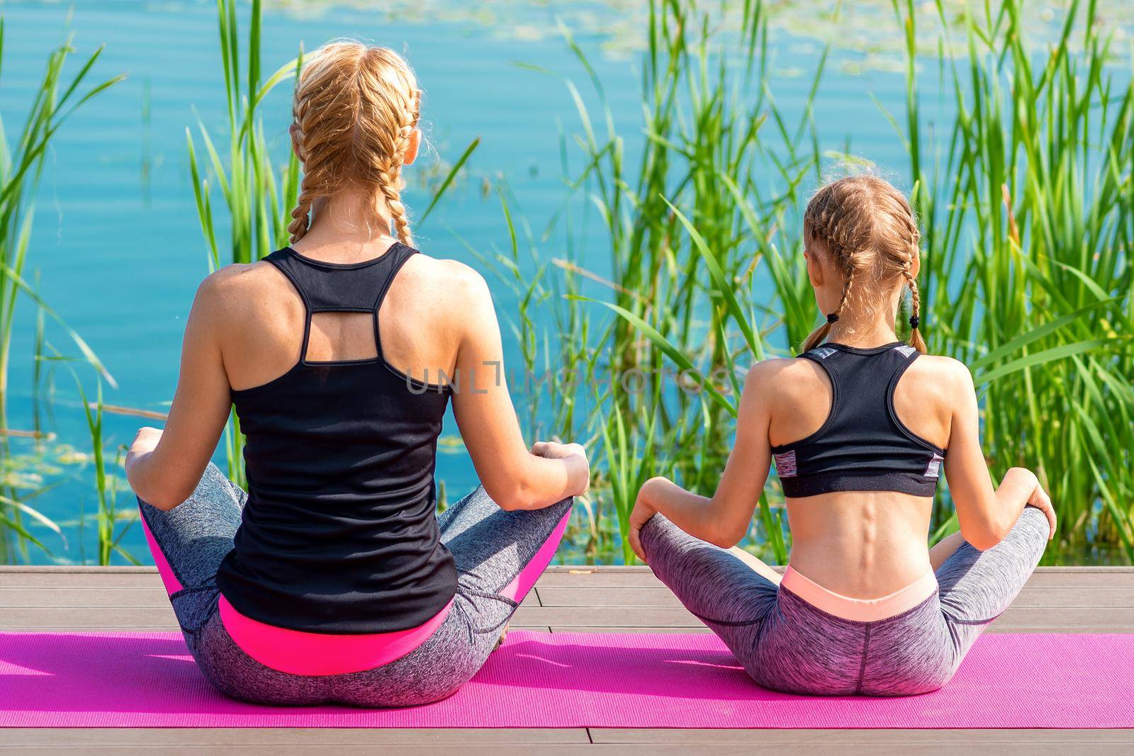 Mother and daughter in sportswear sitting on the mat on the pier near the water outdoor