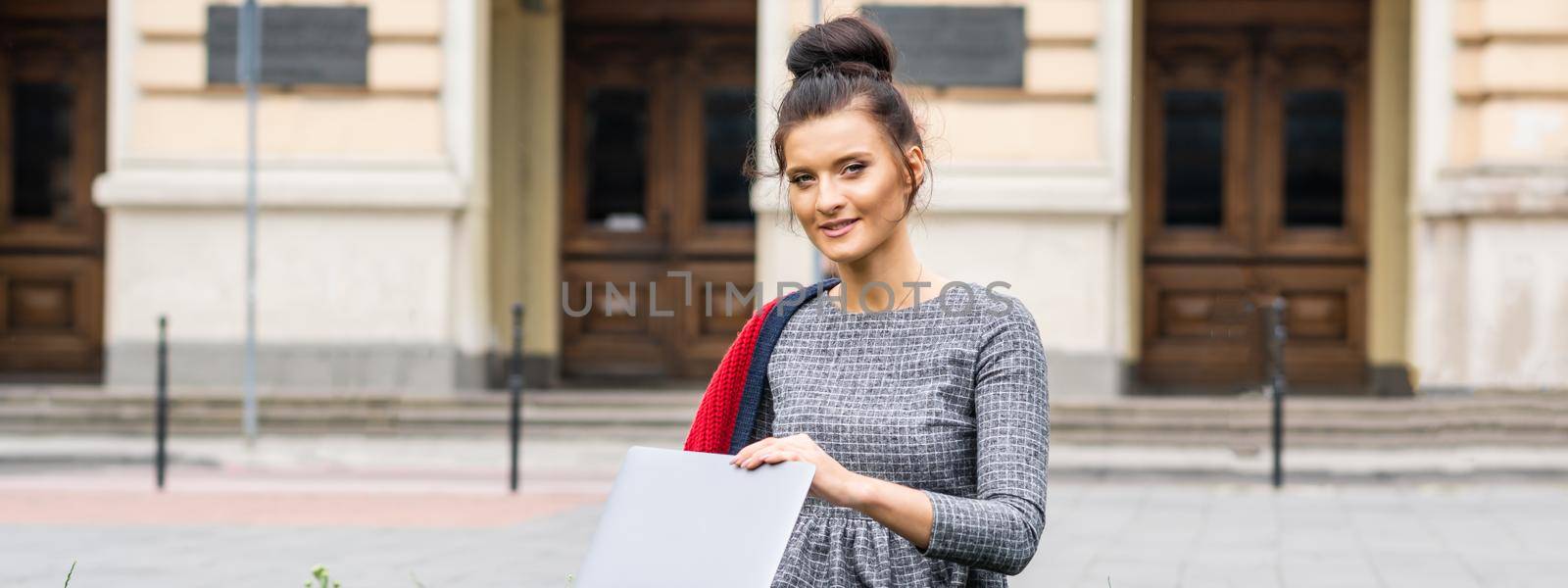 Portrait of a beautiful young smiling student studying with a laptop on the grass near the university building
