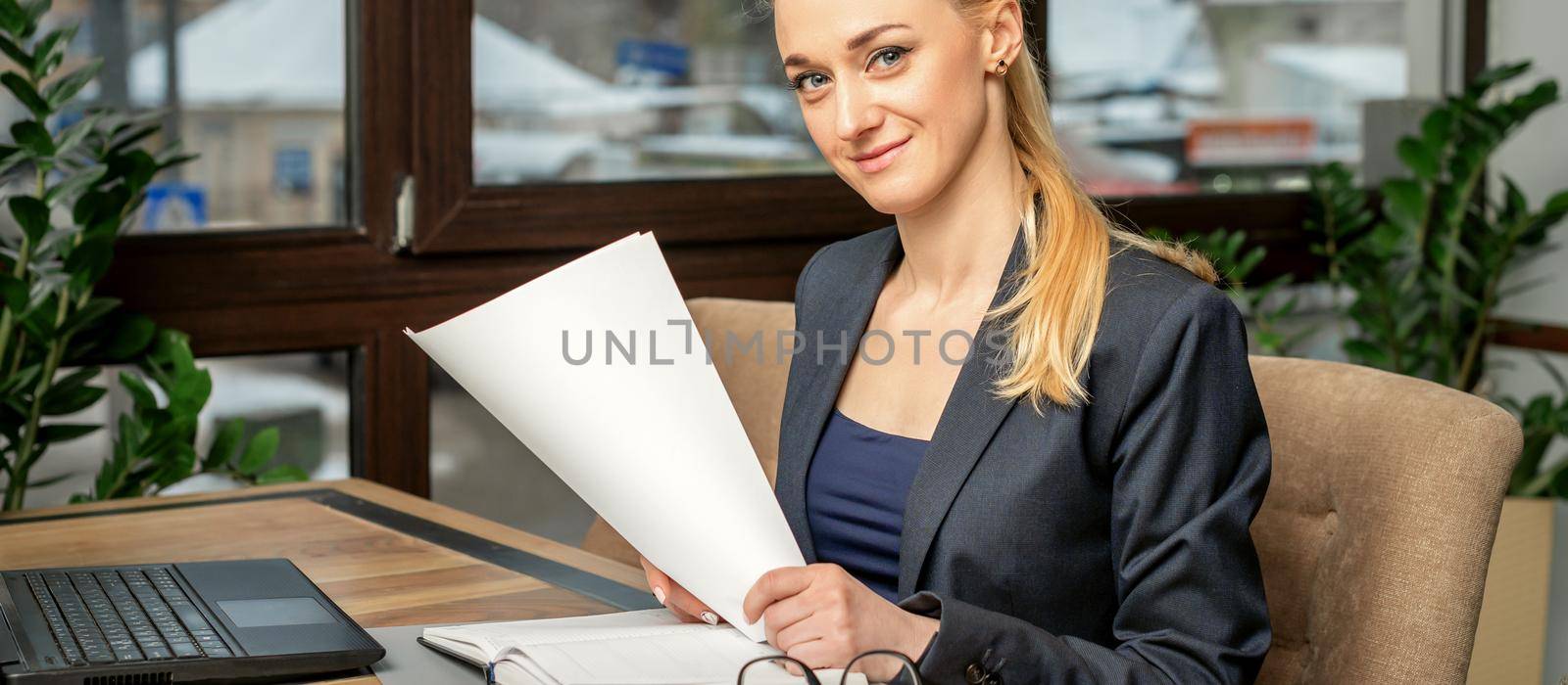 Portrait of a young smiling caucasian businesswoman prepares documents with laptop at the table looking at the camera in cafe
