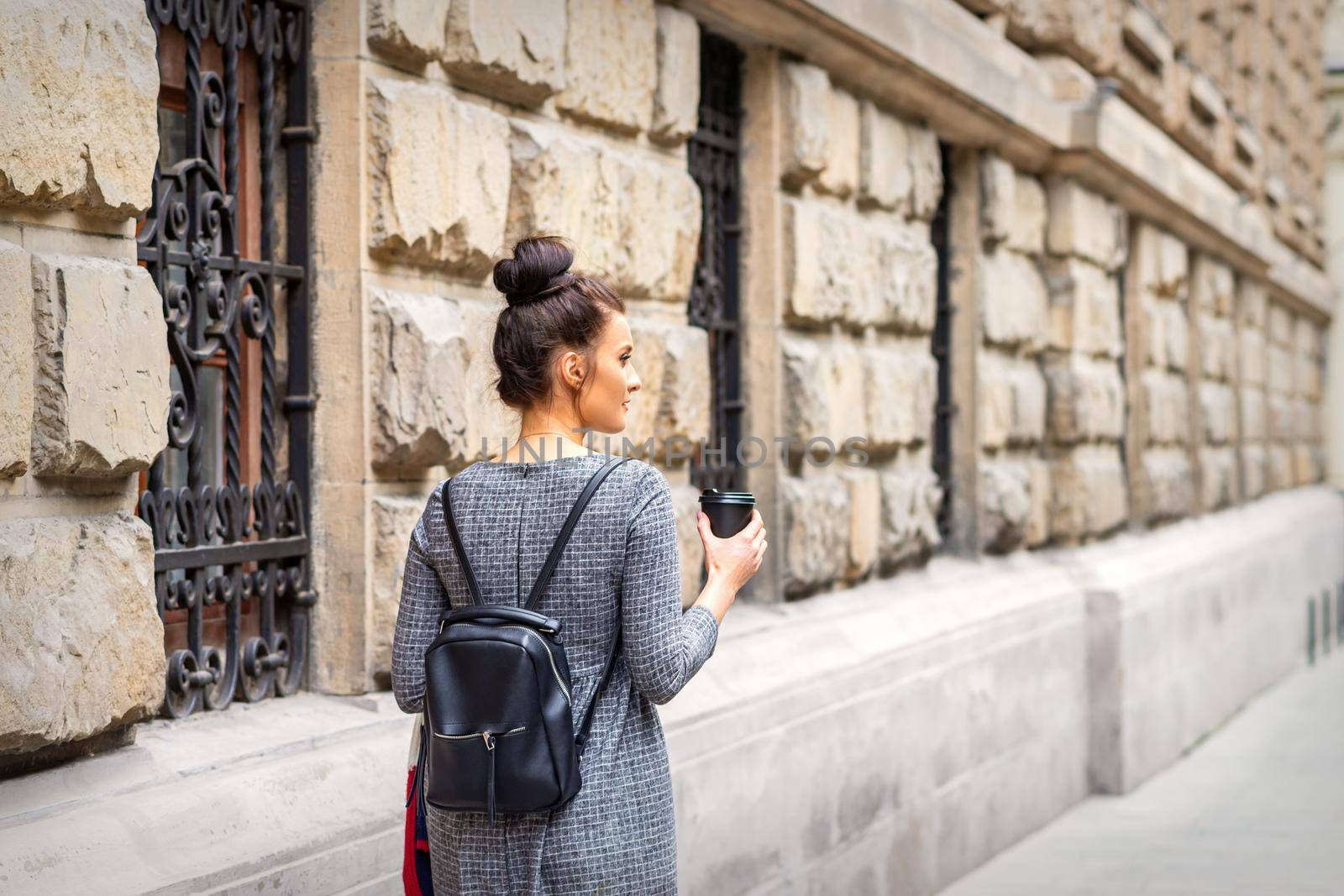 Back view of a young woman with a hot drink and backpack walks in a European city