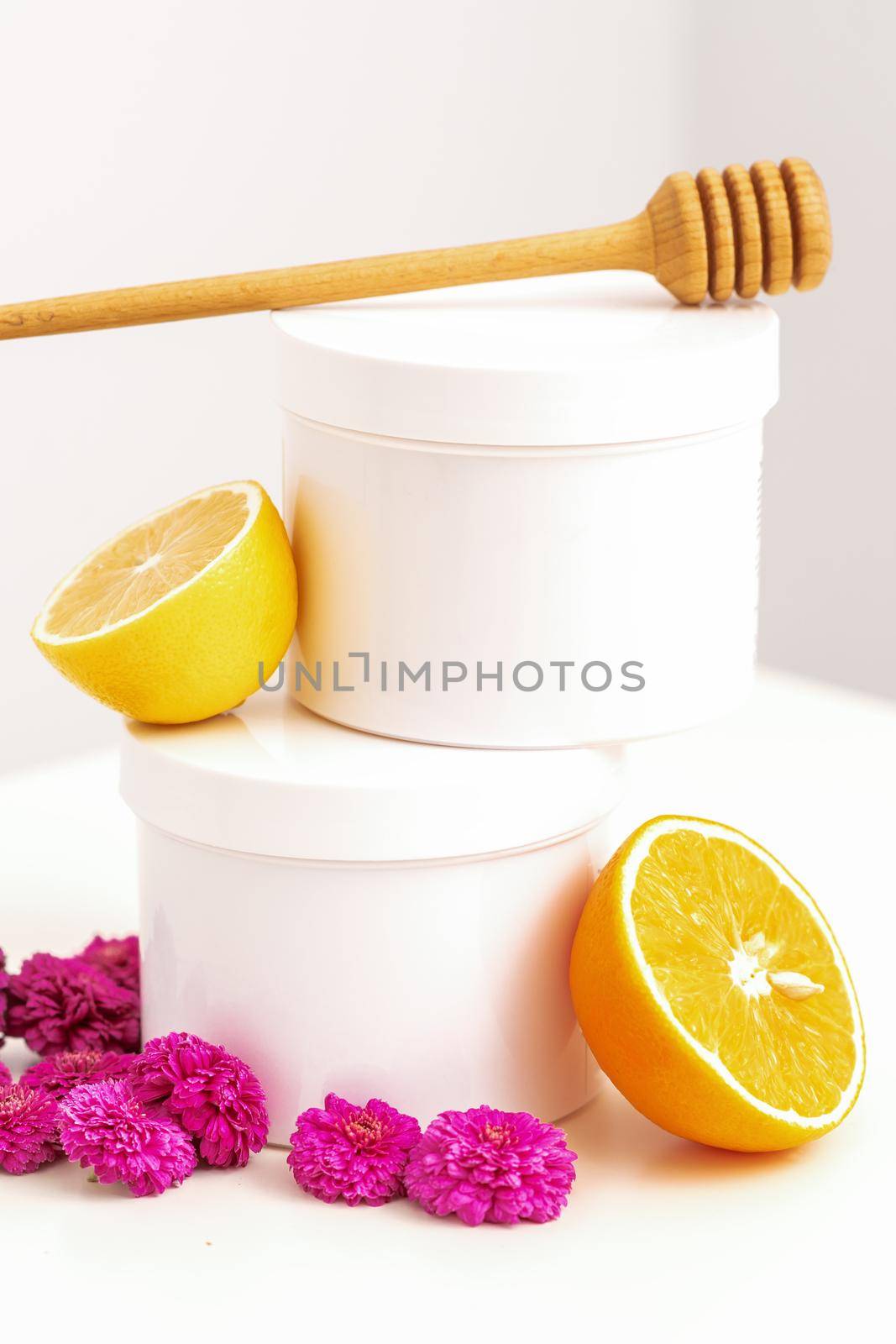 White cosmetic jars with a wooden honey stick, fresh lemon, and chrysanthemums flowers on white background with copy space. Blank space for branding mockup beauty product