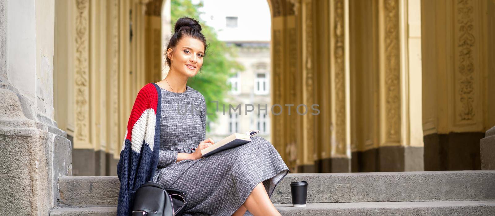Portrait of a young female student with a book sitting on the stairs of building outdoors