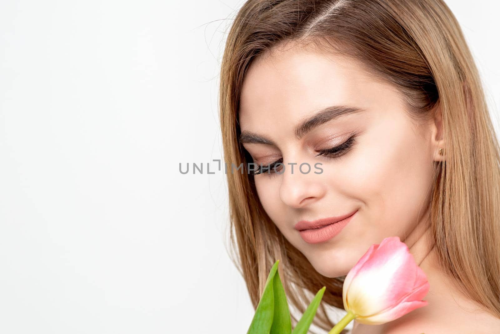 A portrait of a happy young caucasian woman with closed eyes and one pink tulip against a white background with copy space