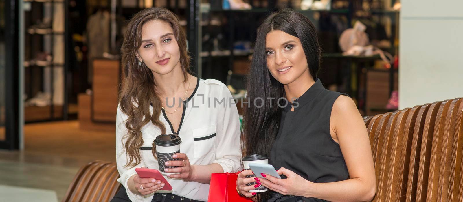 Two beautiful caucasian young women looking at the camera with bags sitting on the bench while resting in the shopping mall