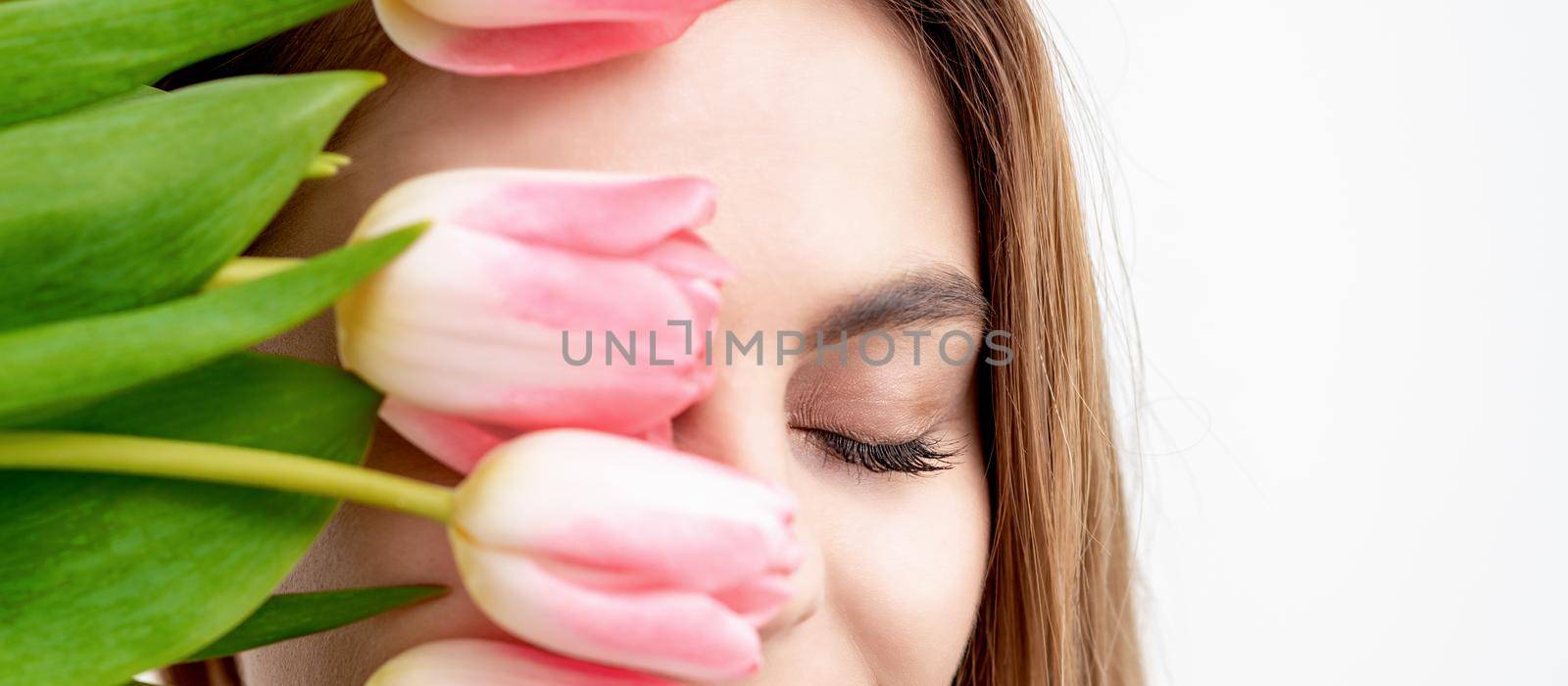 Half face portrait of a happy young caucasian woman with closed eyes and pink tulips cover her face against a white background