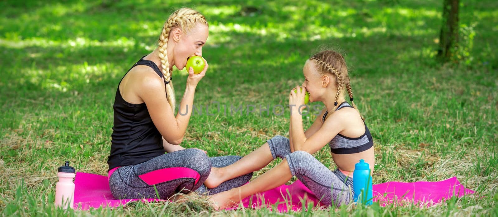 Mother with daughter in sportswear eats apples sitting on a mat in the summer park