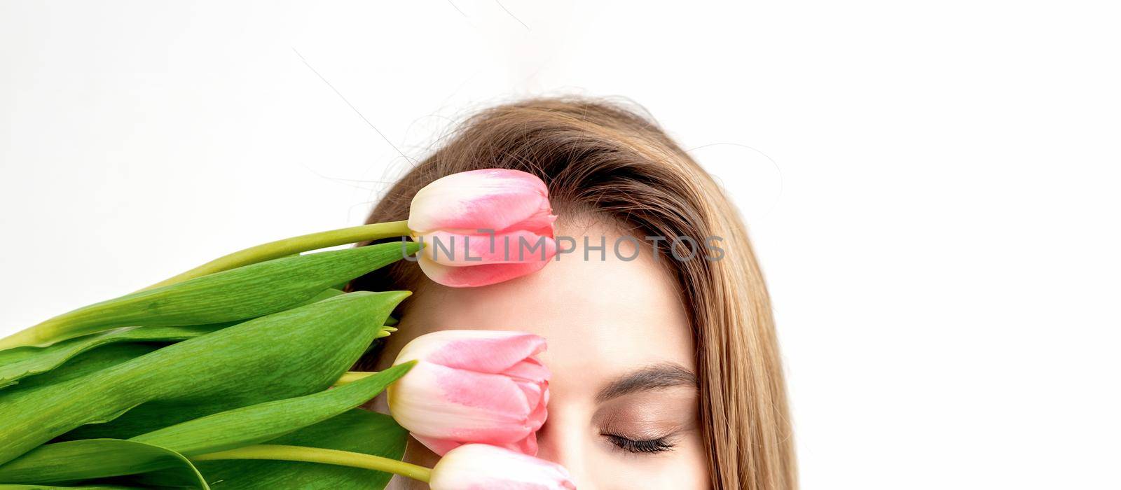 Half face portrait of a happy young caucasian woman with closed eyes and pink tulips cover her face against a white background