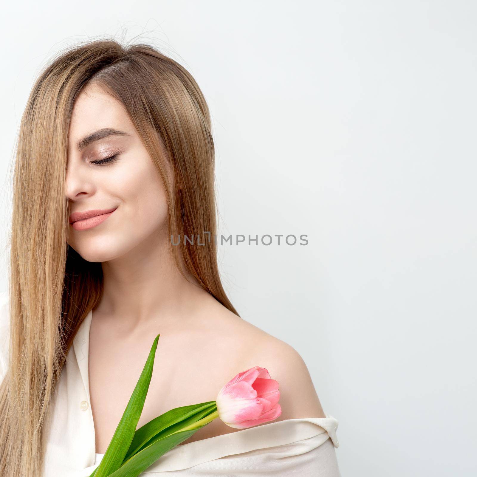 A portrait of a happy young caucasian woman with closed eyes and one pink tulip against a white background with copy space
