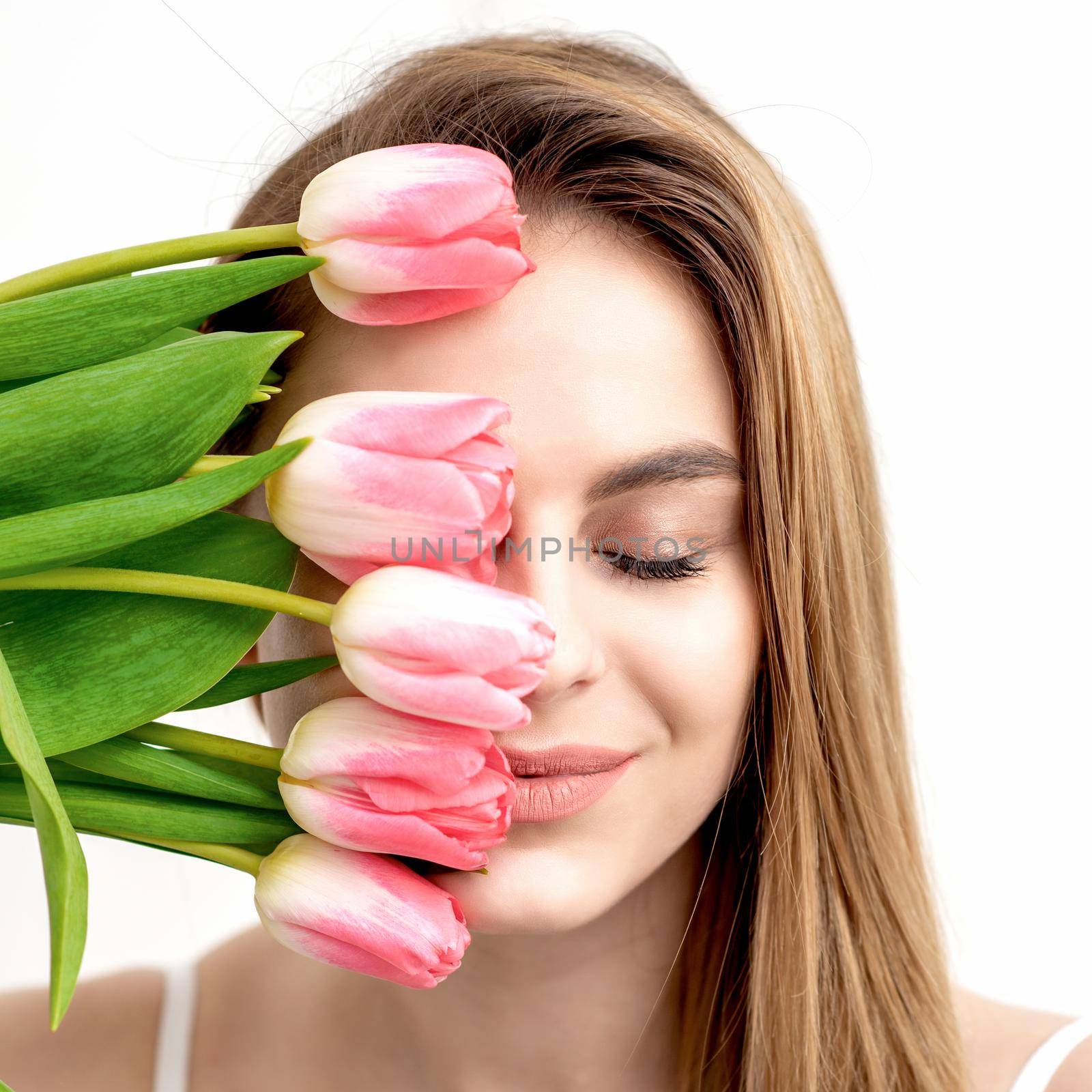 A portrait of a happy young caucasian woman with closed eyes and pink tulips cover her face against a white background
