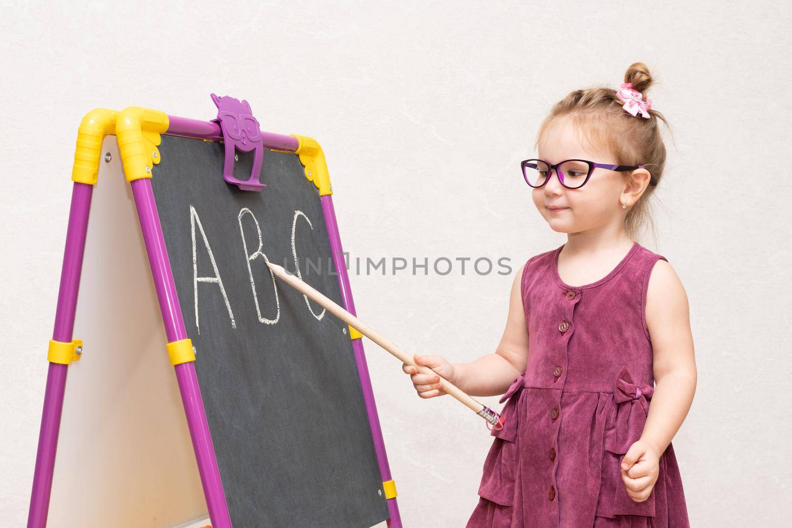 cheerful emotional girl of three years with a strict hairstyle like a teacher in a Burgundy dress and glasses. near the blackboard shows students. the letters in the alphabet....