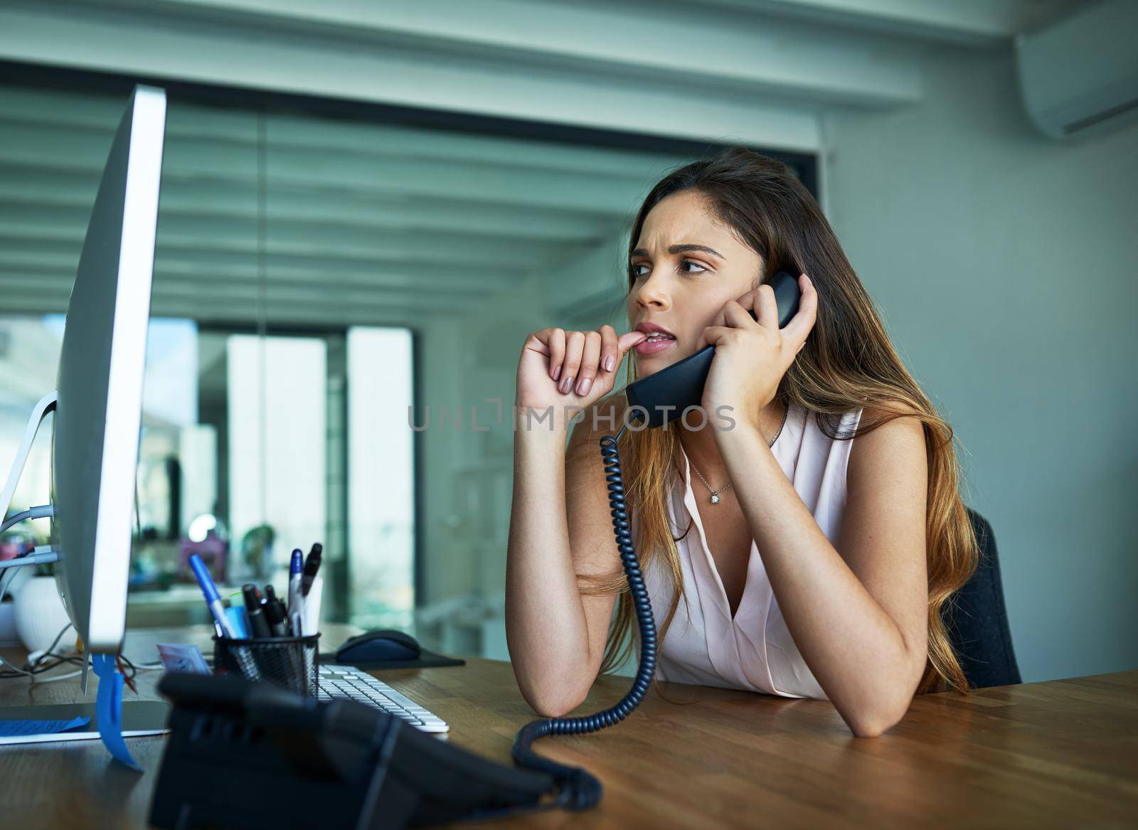 Sometimes anxiety gets the best of her. a young businesswoman biting her nails while talking on a phone in an office. by YuriArcurs