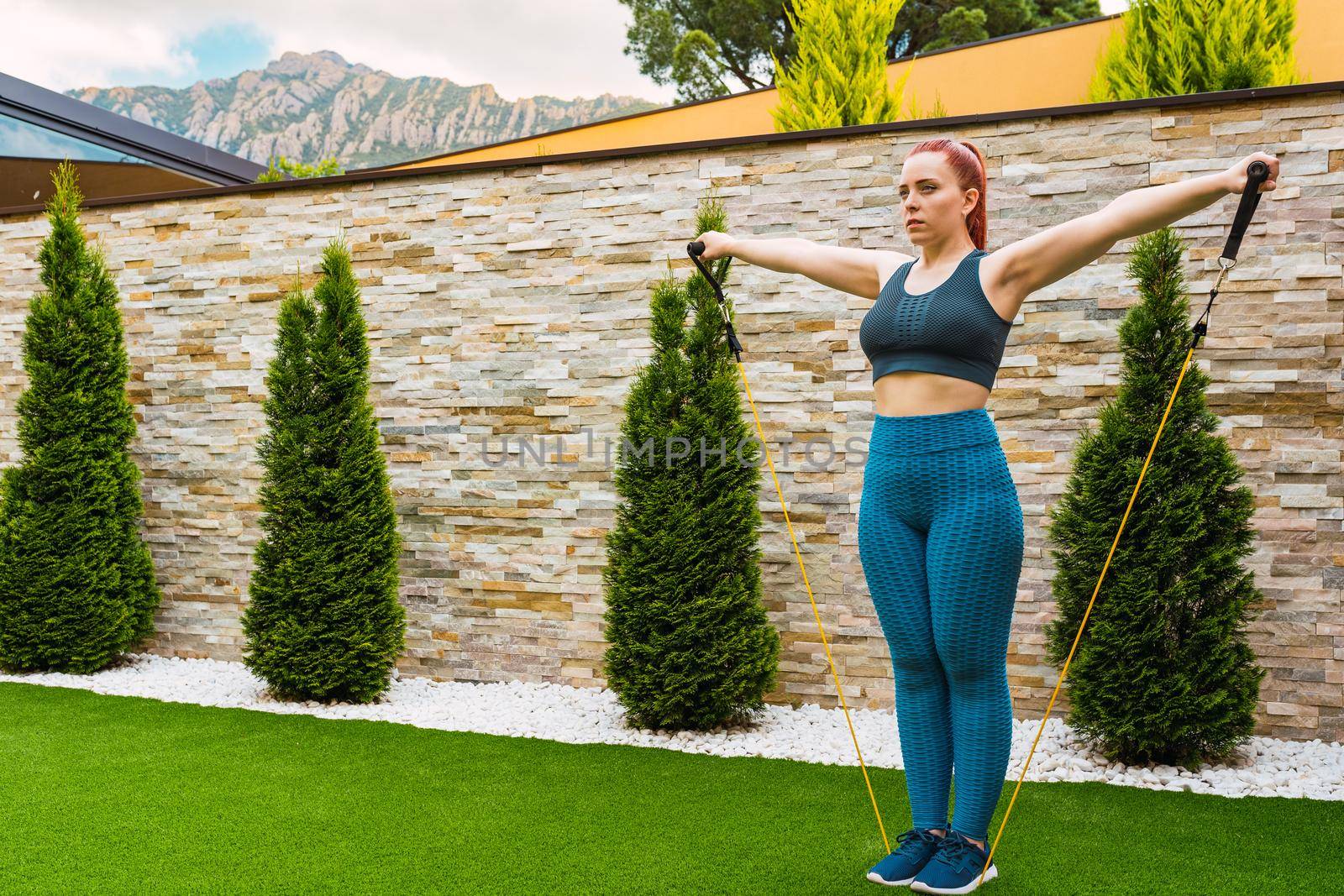 young sportswoman, stretching her body muscles with elastic bands outdoors in the garden of her house. concept of health and well-being. by CatPhotography