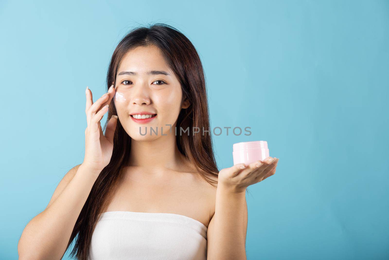 Asian young woman are not very beautiful hold jar of skin cream for face and apply anti-aging moisturizer studio isolated on blue background, Happy female pointing finger her face, pursuit of beauty