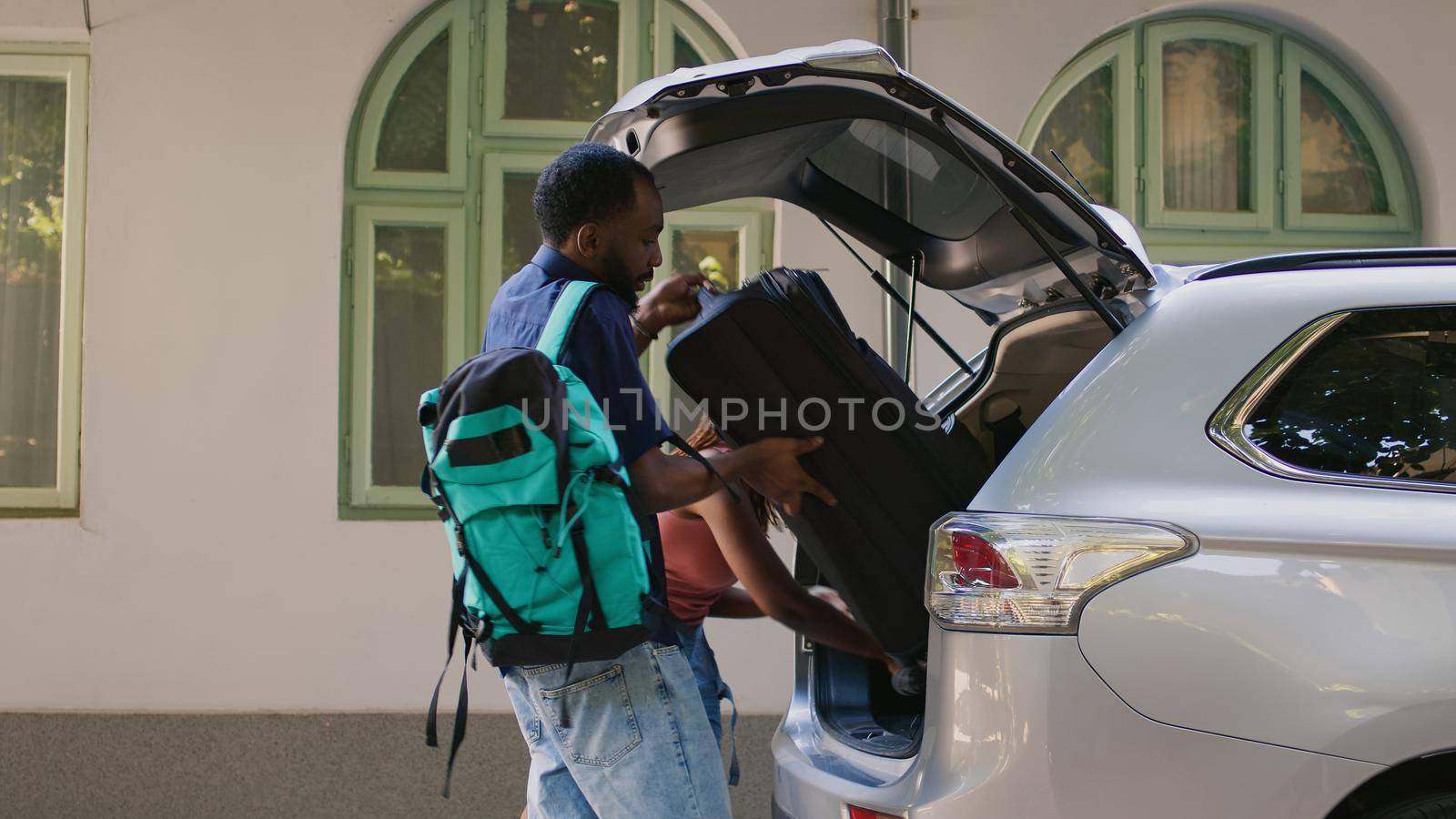 Married couple with luggage going on vacation while loading car trunk with trolley and backpack. Boyfriend and girlfriend getting ready for citybreak departure while putting baggage inside vehicle.