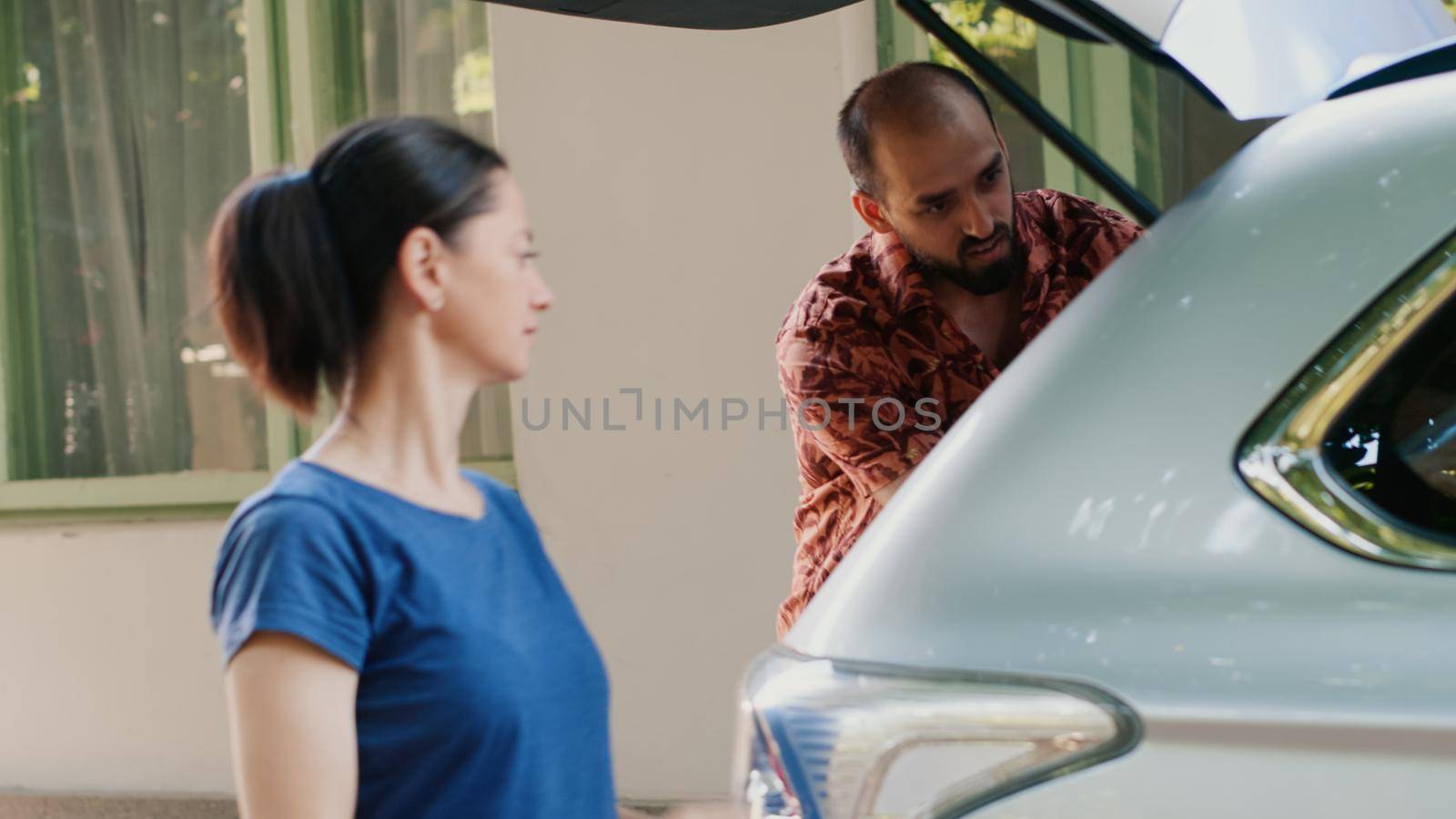 Parents putting voyage luggage and baggage inside car trunk while going on summer holiday field trip. Casual family going on summer cityscape tour while loading trolleys inside vehicle.