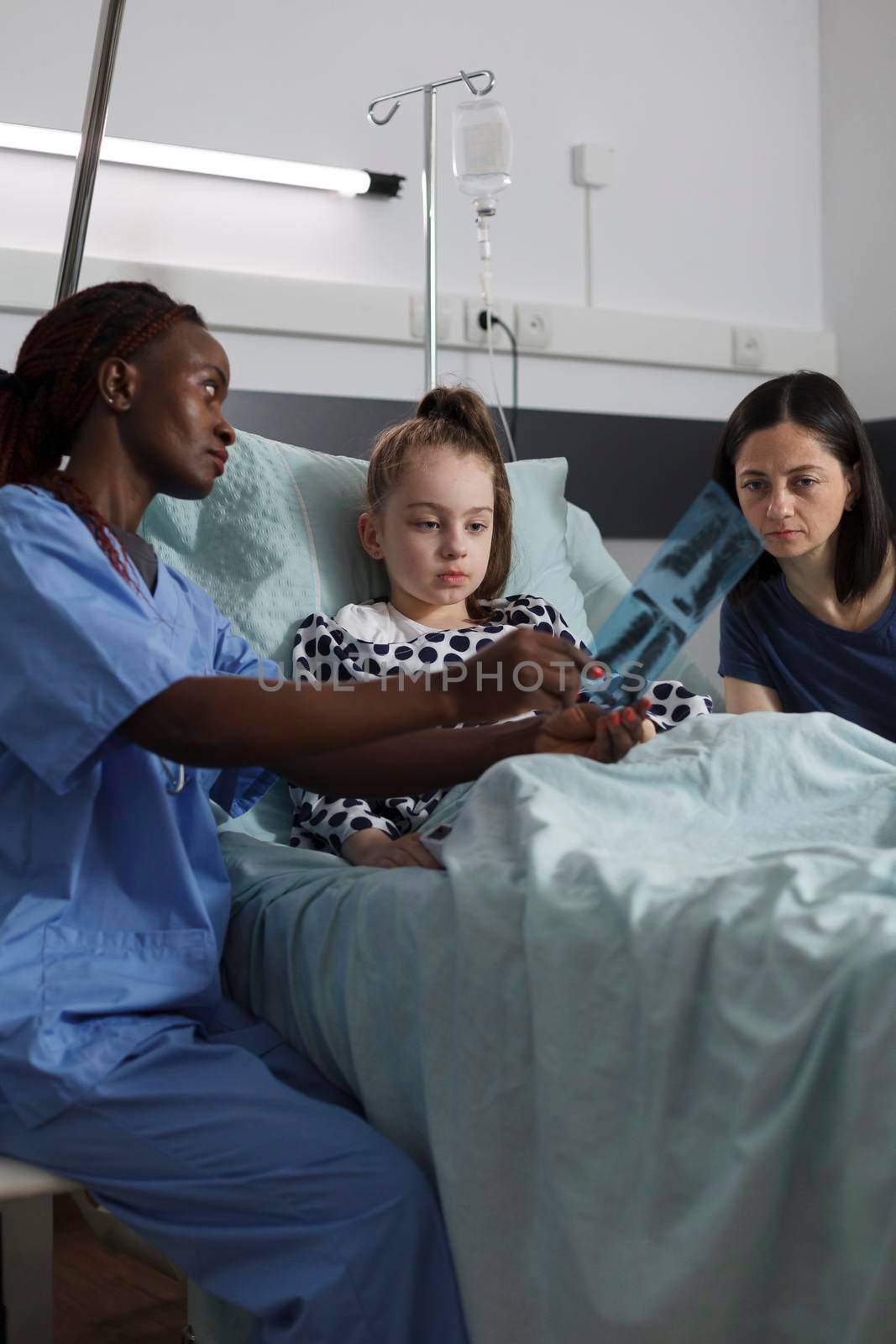 African american nurse showing radiography scan image to sick girl while mother sitting beside her. Pediatric healthcare facility staff analyzing MRI results of ill kid under treatment resting