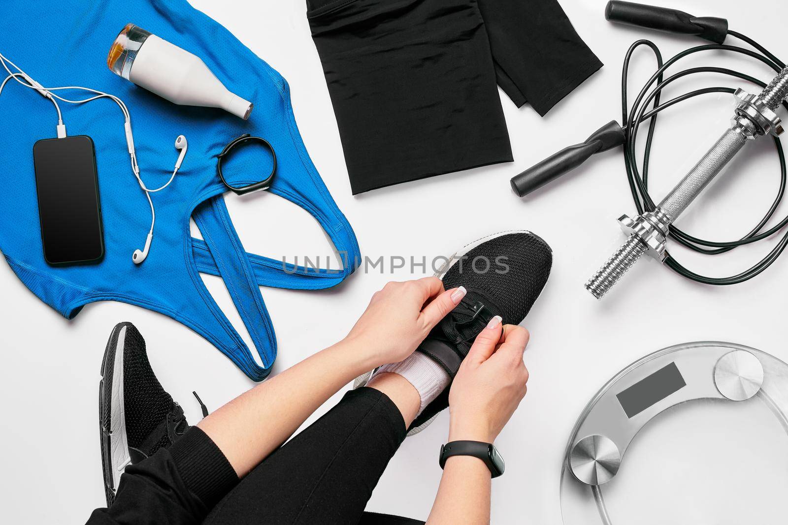 Young woman in sporting leggings laces sneakers, preparing for training. Accessories for sports, bottle of water, smartphone, headphones, fitness bracelet, dumbbells on white background flat lay top view. Fitness concept