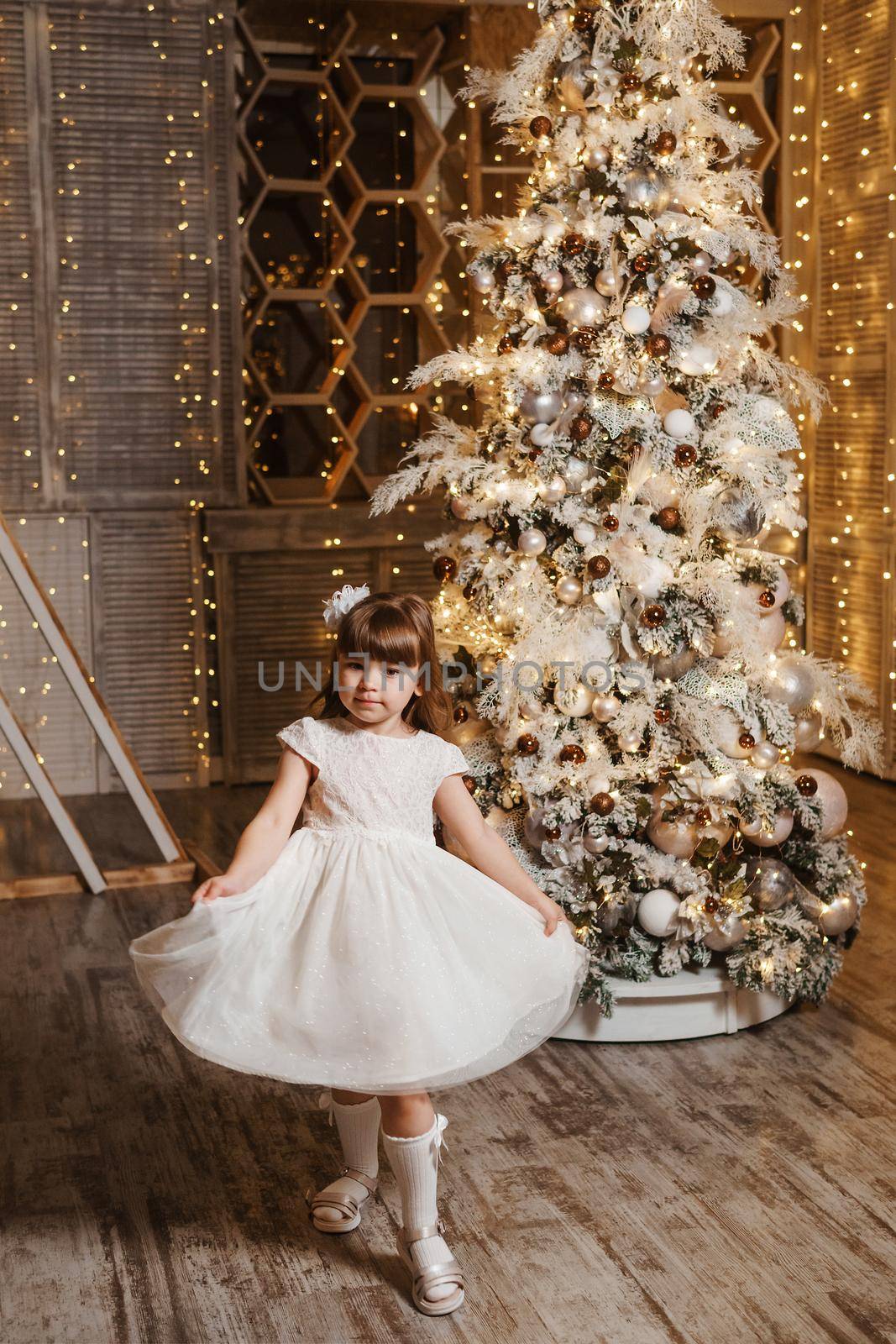 A girl in a festive light dress next to a Christmas tree, lights of garlands in the background. The concept of New Year holidays by Annu1tochka