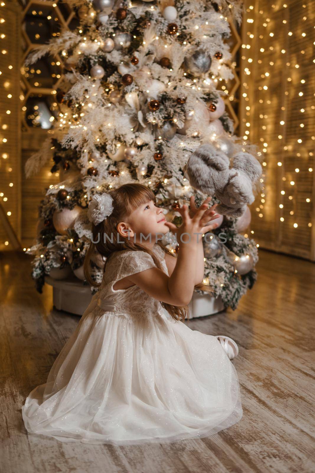 A girl in a festive light dress next to a Christmas tree, lights of garlands in the background. The concept of New Year holidays.