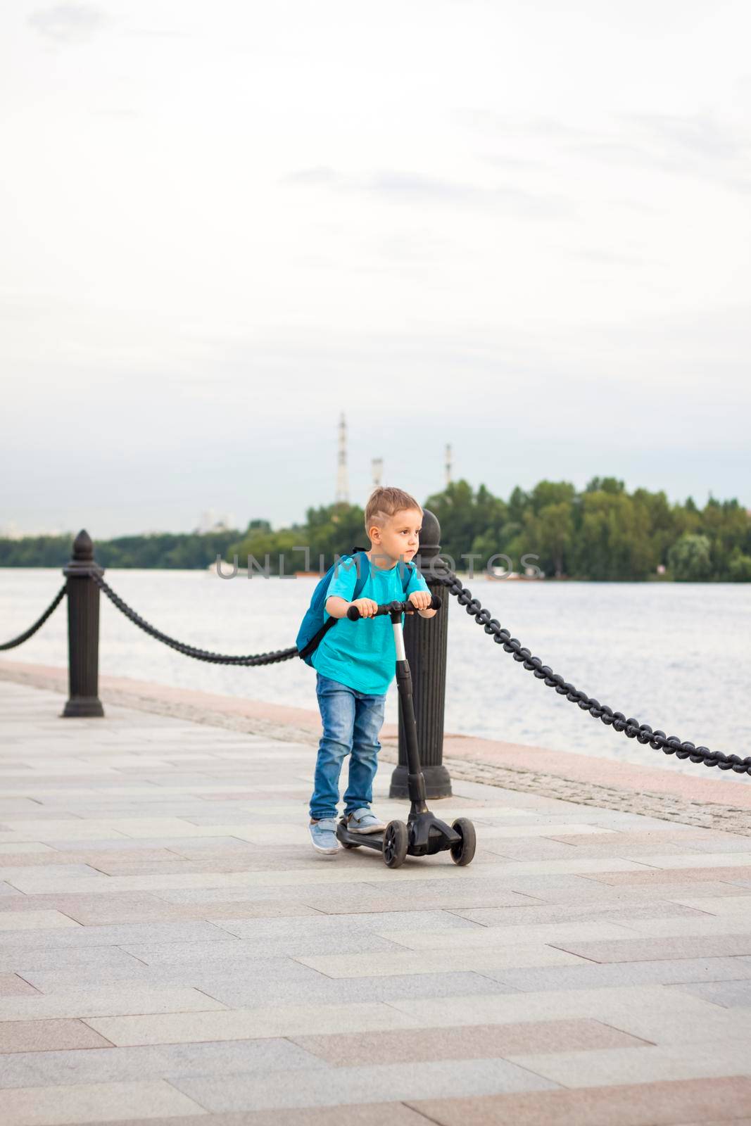 A boy on a scooter along the embankment of the city. Journey. Backpack on the back. The face expresses natural joyful emotions. Not staged photos from life.