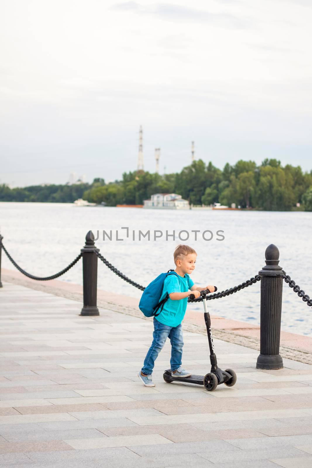 A boy on a scooter along the embankment of the city. Journey. Backpack on the back. The face expresses natural joyful emotions. Not staged photos from life.