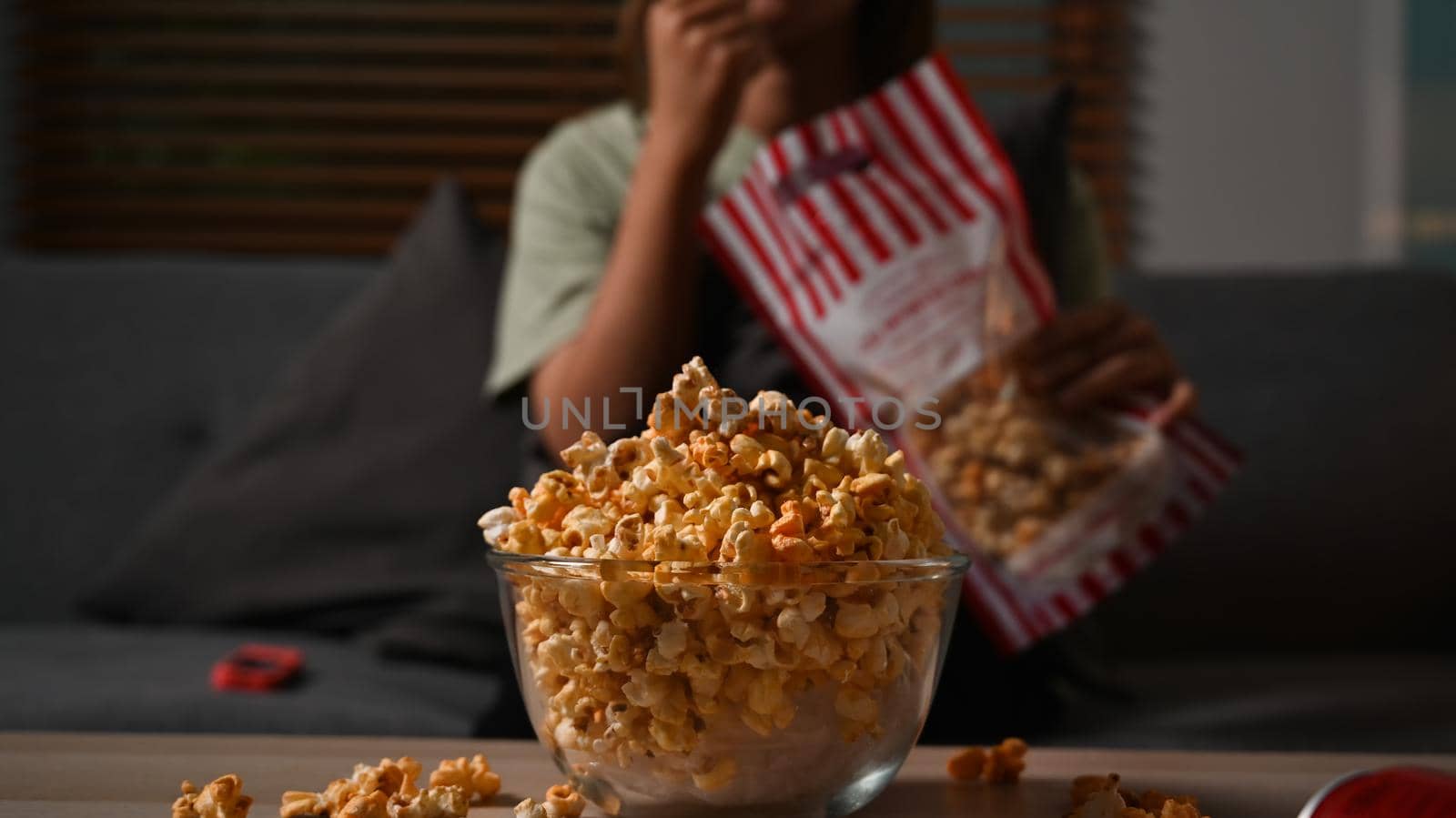 Bowl of popcorn and on wooden table with young woman watching television on couch in background. Relaxation and hobby concept.