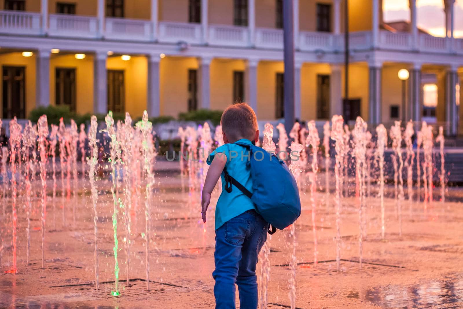 Children have fun frolicking in the fountains near the building in the park in the sunset light. Evening walk around the city