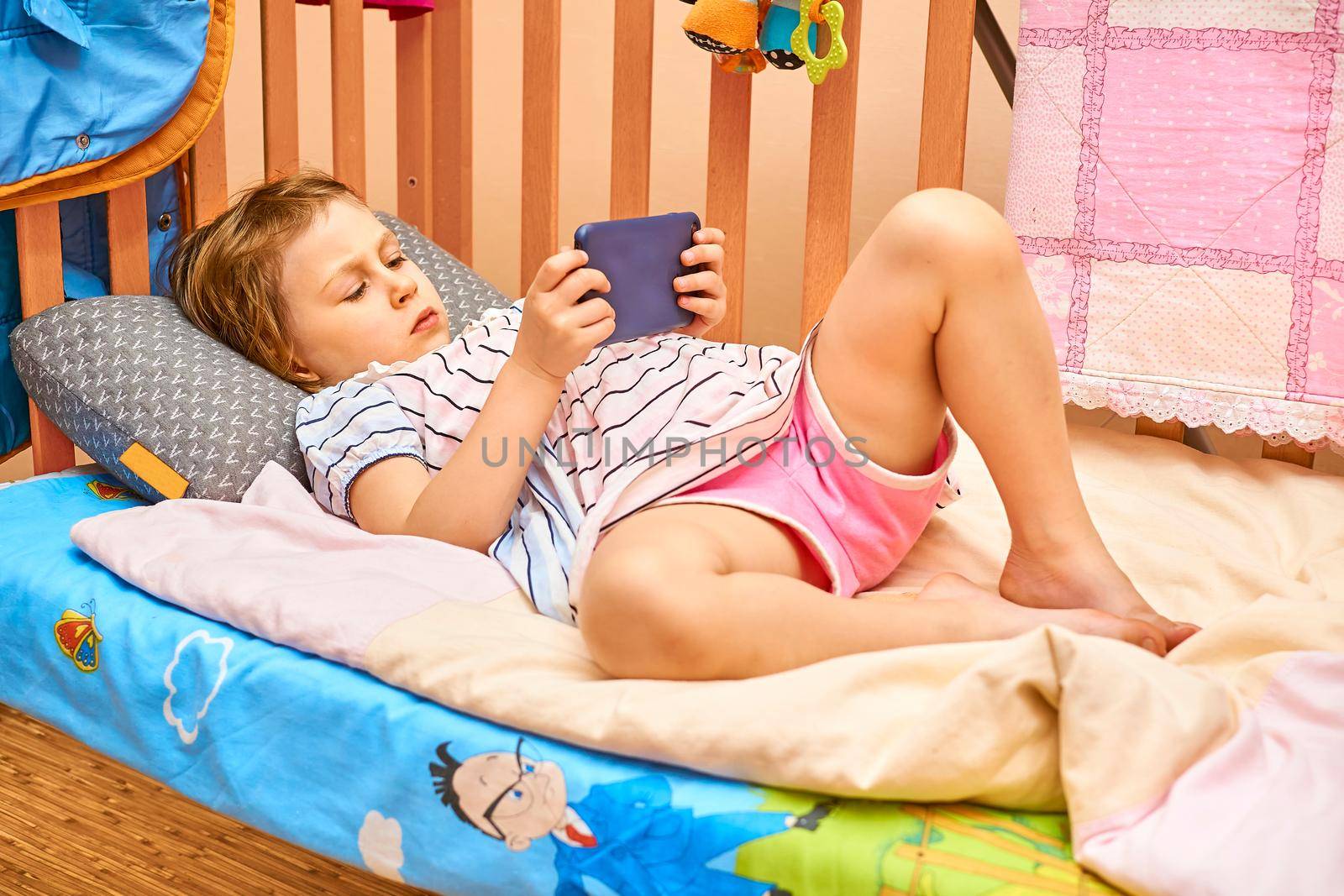 a young human being below the age of puberty or below the legal age of majority. A cute child lying in a crib looking news, games, fairy tales on a smartphone.