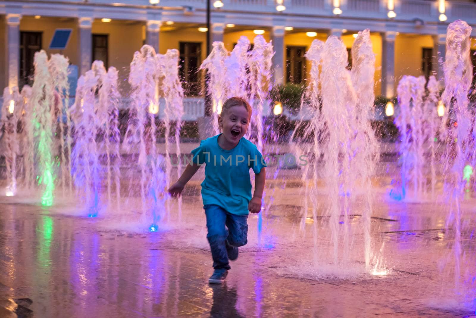 Children have fun frolicking in the fountains near the building in the park in the sunset light. Evening walk around the city