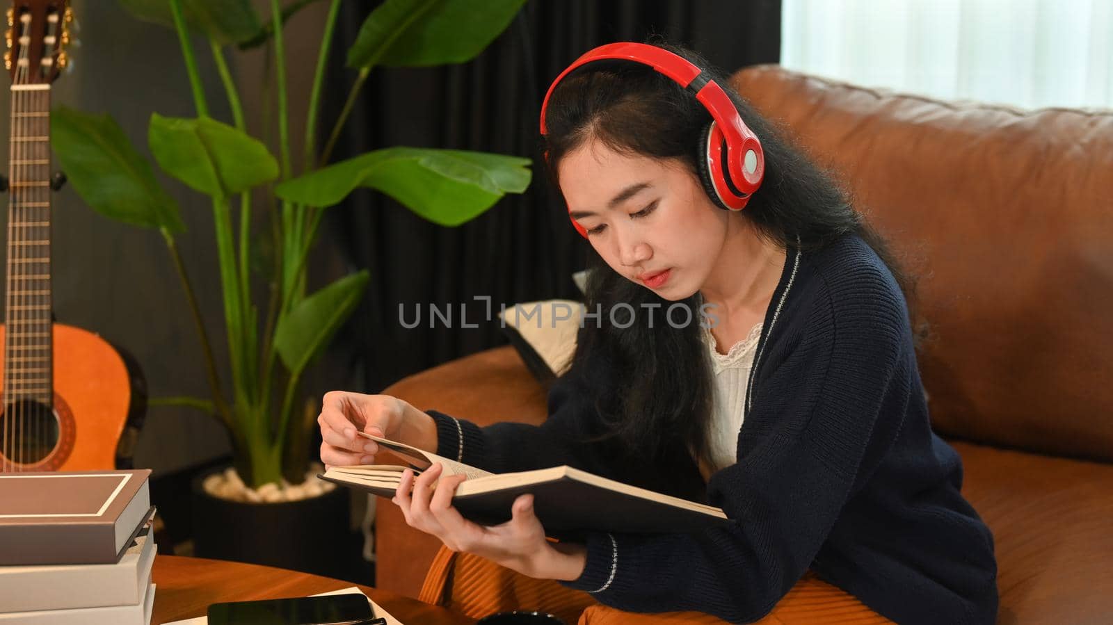 Beautiful young woman listening to music on headphone and reading book, spending leisure time in cozy winter or autumn weekend at home.