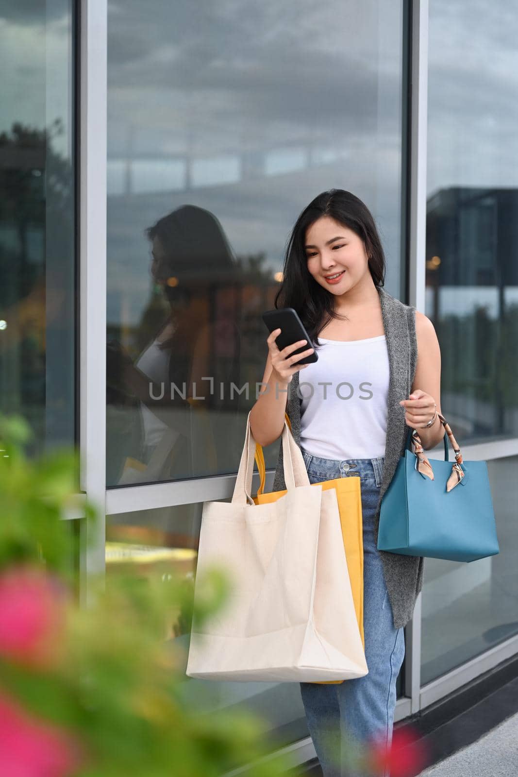 Portrait of young woman with shopping bags using smart phone and walking in the city street by prathanchorruangsak