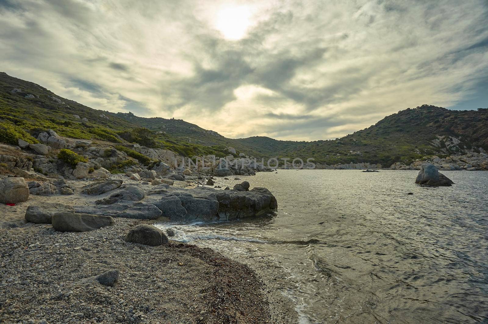 Seascape at sunset containing a magnificent Mediterranean beach with rocky headlands and mountains under a cloudy sky with glimmers of sunshine.