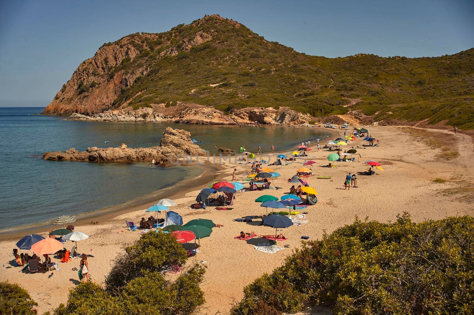 Beach of southern Sardinia (Cala Sa Figu) inserted in a natural bay full of tourists with umbrellas on vacation.