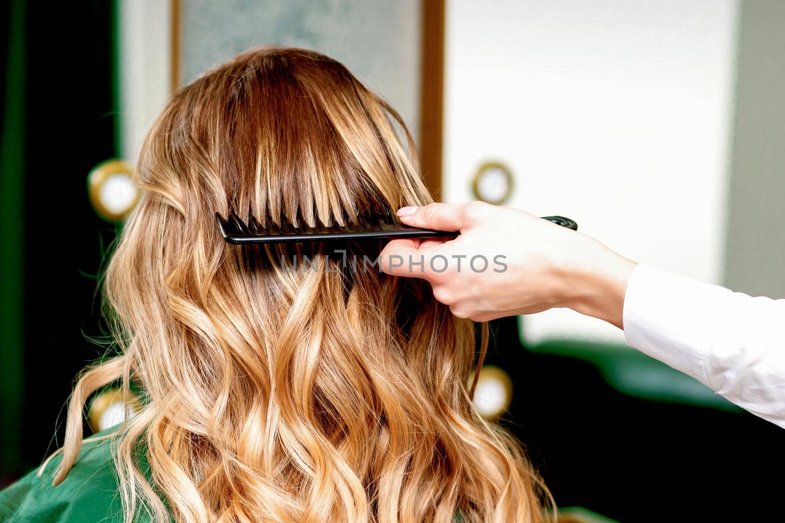 Back view of hairdresser combing wavy hair of a young blonde woman in a beauty salon