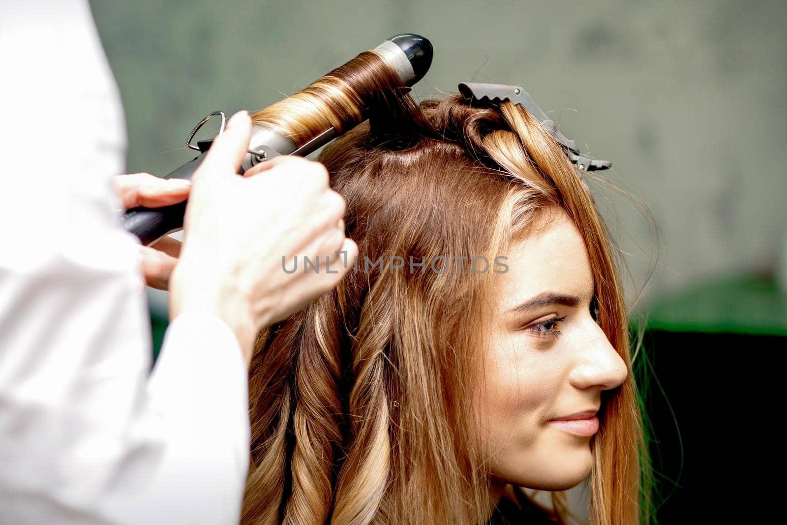 Professional hairdresser makes curls with a curling iron for a young woman with long red hair in a beauty salon