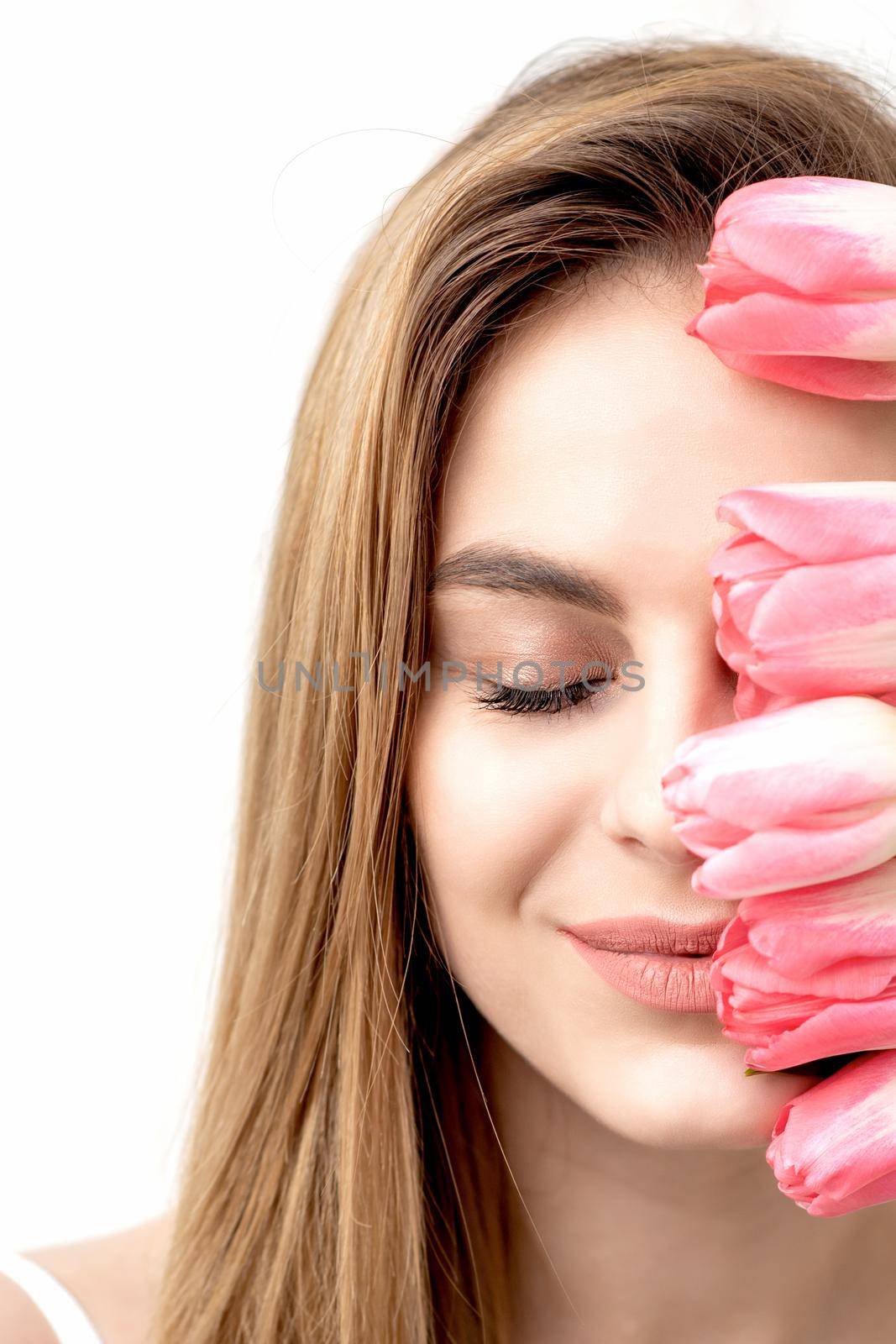 A portrait of a happy young caucasian woman with closed eyes and pink tulips cover her face against a white background