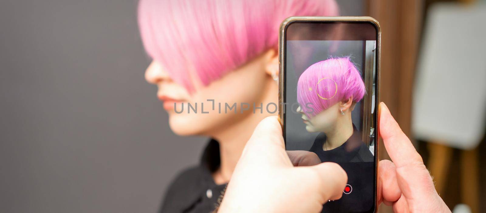 The hairdresser takes pictures of the short pink hairstyle of a young woman on the smartphone