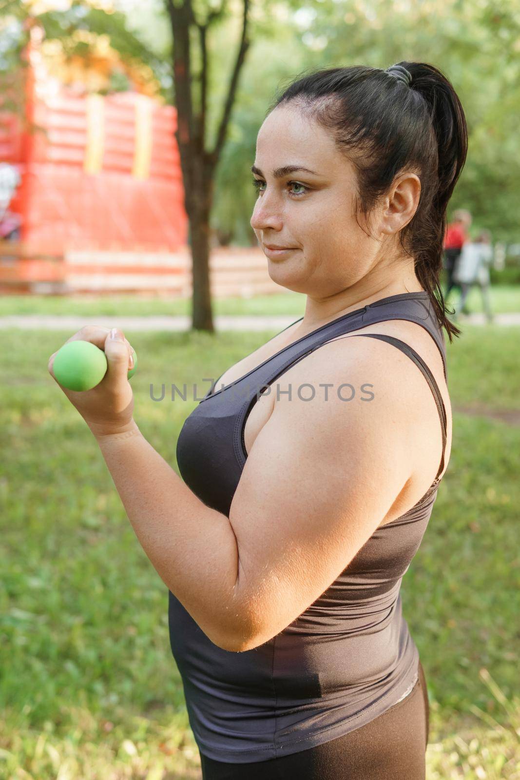 A charming brunette woman plus-size body positive practices sports in nature. Exercises with dumbbells.