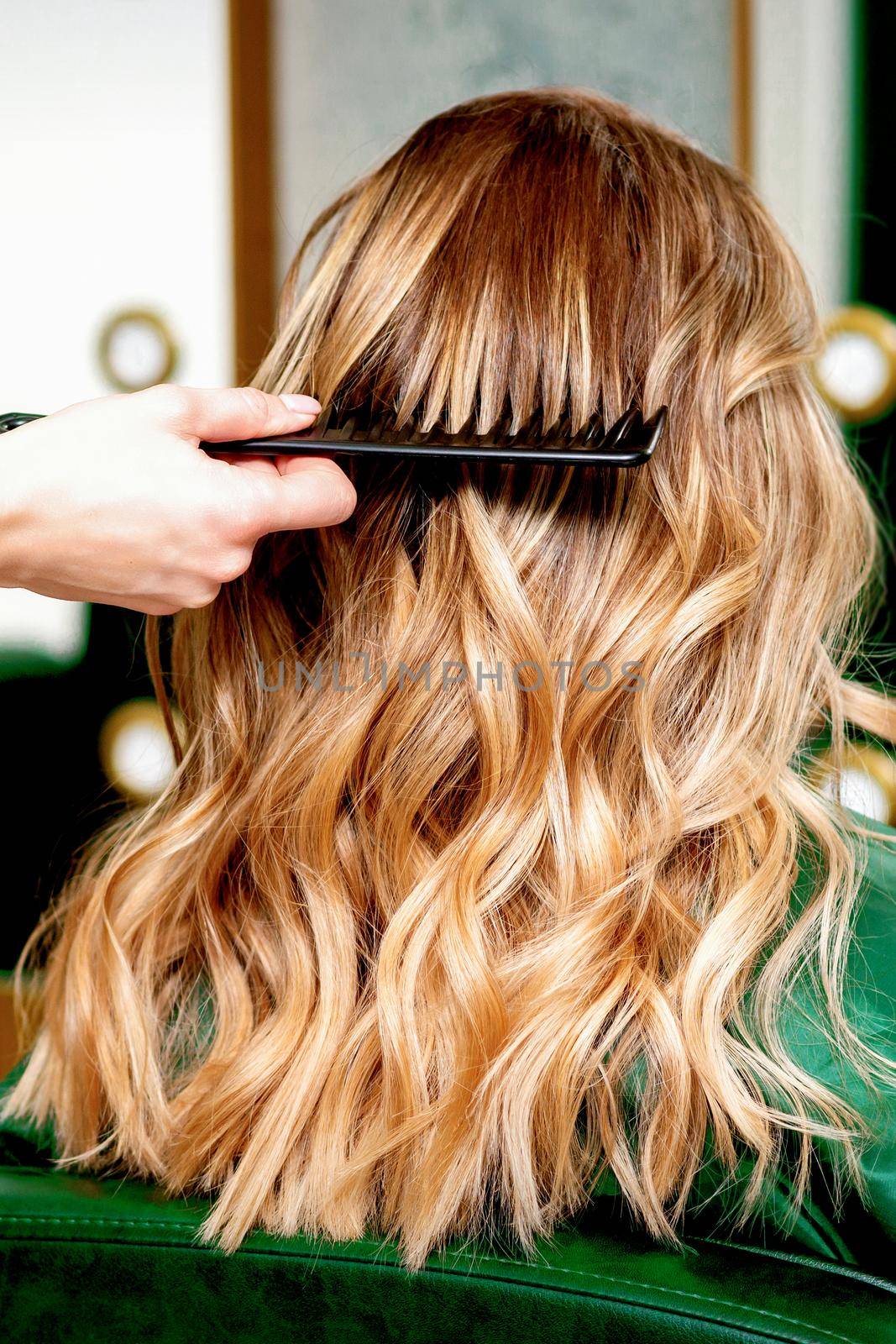 Back view of hairdresser combing wavy hair of a young blonde woman in a beauty salon