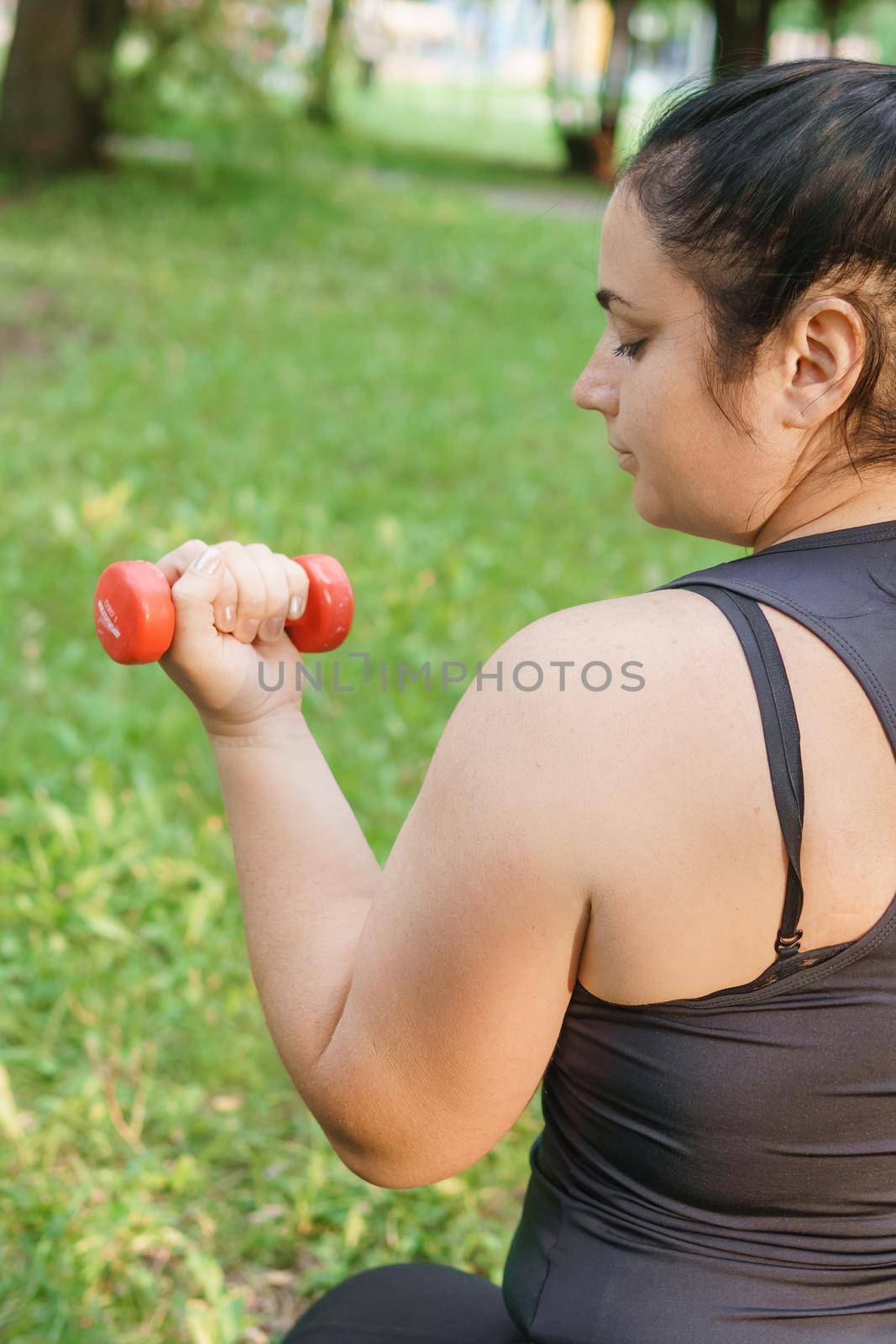 A charming brunette woman plus-size body positive practices sports in nature. Exercises with dumbbells.